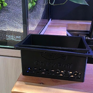 May include: Black rectangular container with the text "Oh My Puff" and a fish logo. The container has a series of hexagonal cutouts and sits on a wooden surface. A fish tank with plants is in the background.