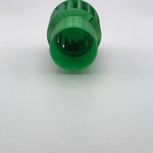 May include: A bright green, cylindrical object with a ridged exterior and a hollow interior. The object has a series of vertical slots around its circumference. The item appears to be made of plastic and is set against a white background.