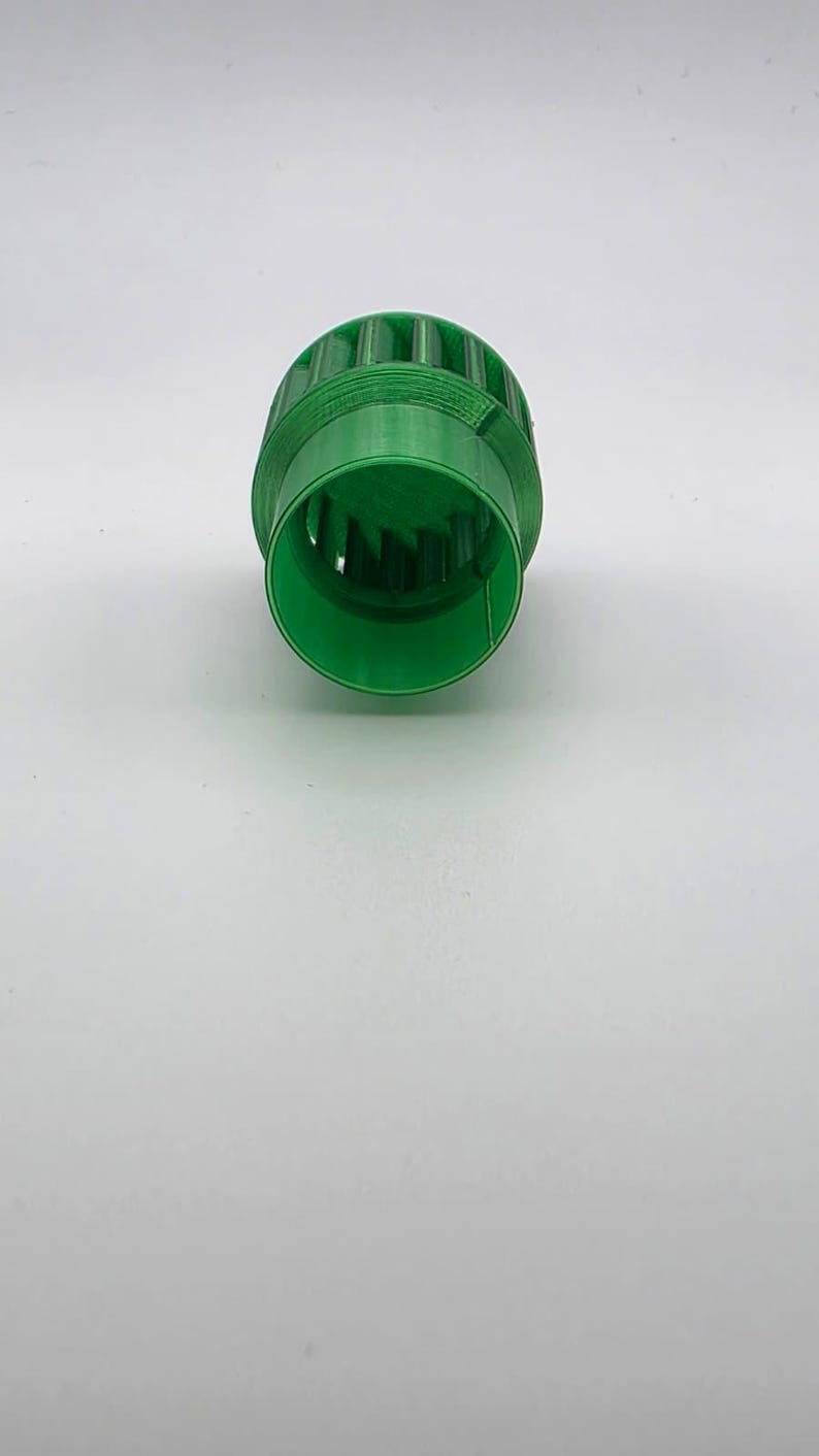 May include: A bright green, cylindrical object with a ridged exterior and a hollow interior. The object has a series of vertical slots around its circumference. The item appears to be made of plastic and is set against a white background.