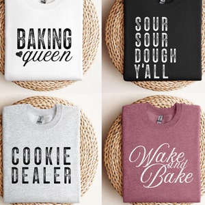 以下が含まれることがあります： 焼き菓子をテーマにしたテキストが書かれた、異なる4つのTシャツ。最初のTシャツは白で、黒い文字で「Baking queen」と書かれている。2番目のTシャツは黒で、白い文字で「Sour Sour Dough Y'all」と書かれている。3番目のTシャツはグレーで、黒い文字で「Cookie Dealer」と書かれている。4番目のTシャツはピンクで、白い文字で「Wake and Bake」と書かれている。