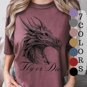 May include: A maroon t-shirt with a black and white illustration of a dragon's head and the text "Fly or Die". The shirt is available in 7 colors.