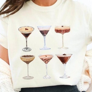 Peut inclure: Un t-shirt blanc avec un motif de six cocktails différents. Les cocktails sont tous dans des verres à martini et ont des liquides et des garnitures de différentes couleurs.