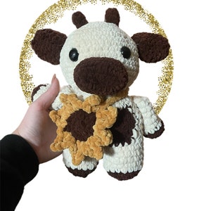 May include: A handmade crochet plush toy shaped like a cow with cream and dark brown yarn. The cow features black plastic eyes, small horns, and a dark brown snout. It wears a crocheted sunflower accessory around its neck with yellow petals.
