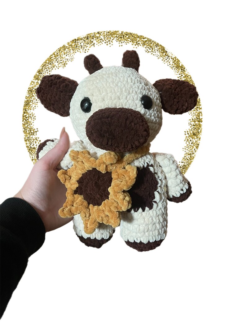 May include: A handmade crochet plush toy shaped like a cow with cream and dark brown yarn. The cow features black plastic eyes, small horns, and a dark brown snout. It wears a crocheted sunflower accessory around its neck with yellow petals.