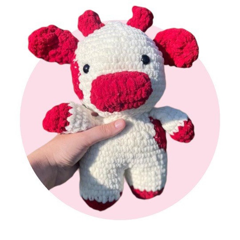 May include: A handmade crochet plush toy shaped like a cow, featuring a cream-colored body with bright red patches on the ears, horns, snout, and feet. The stuffed animal has two small black plastic eyes and a soft, textured yarn construction.