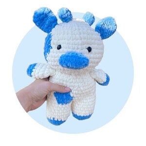 May include: A plush stuffed animal crafted from white and blue yarn in a crochet style. The creature features a rounded head with two blue ears, two small horns, a blue snout, and two black bead eyes. It has a white body with blue patches.