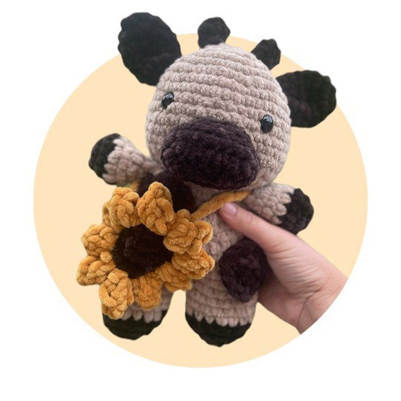 May include: A crochet plush toy shaped like a cow with light brown and dark brown yarn. The cow features dark brown ears, hooves, and a snout, with a yellow and brown sunflower accessory attached to its body by a thin yarn strap.