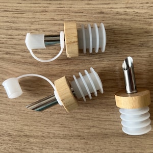 Może przedstawiać: Trzy drewniane korki do butelek z białymi silikonowymi pierścieniami i stalowymi dyszami. Dwa korki mają pętlę do zawieszenia. Jedna dysza ma spiczasty koniec.