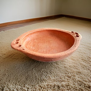 Può includere: Una ciotola in terracotta di forma arrotondata con un bordo largo. La ciotola ha una superficie liscia di colore marrone rossiccio e quattro piccoli fori sul lato.