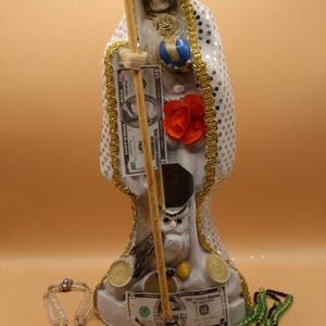 May include: A white statue of Santa Muerte, the folk saint of death, holding a scythe and a globe. The statue is adorned with gold trim, a red rose, and dollar bills. Various rosaries and coins are at the base.