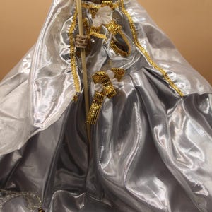 Puede incluir: Una figura decorativa de esqueleto con una túnica plateada con adornos dorados y sosteniendo un bastón de madera con una pequeña guadaña. La figura está adornada con detalles dorados y joyas.