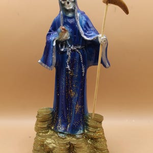 Könnte beinhalten: Eine blau-silberne Statue einer bekleideten Skelettfigur, die eine Sense hält. Die Figur steht auf einem Haufen Goldmünzen. Ein silberner Schlüsselanhänger mit einem kleinen Anhänger befindet sich im Vordergrund.