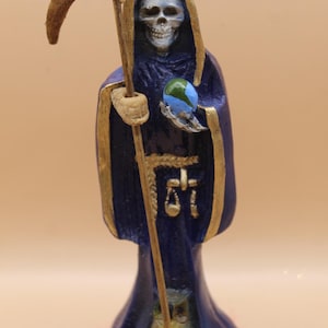 Könnte beinhalten: Eine Figur eines Skeletts in einem dunkelblauen Gewand, das eine Sense und einen Globus hält. Die Figur hat goldfarbene Verzierungen, und der Globus ist blau und grün. Die Figur hat ein detailliertes, schädelartiges Gesicht.