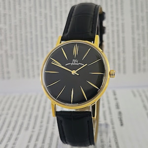 Könnte beinhalten: Eine goldfarbene Armbanduhr mit schwarzem Zifferblatt und goldenen Zeigern. Die Uhr hat ein schwarzes Lederarmband und das Zifferblatt zeigt den Text "ЛУЧ 23 КАМНЯ". Die Uhr ist vor einem verschwommenen Hintergrund platziert.