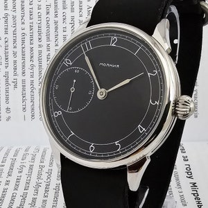 Puede incluir: Un reloj de pulsera plateado con una esfera negra y agujas blancas. La esfera del reloj tiene un pequeño subdial en la posición de las 12 en punto. El reloj está etiquetado como "Молния", que en ruso significa "Relámpago".