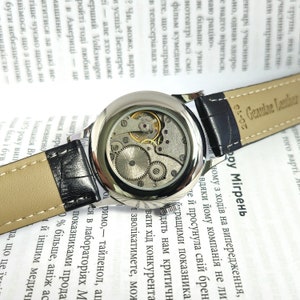Op de afbeelding: Een zilveren polshorloge met een zwarte leren band. De wijzerplaat van het horloge is transparant en laat het innerlijk van het horloge zien. De band is gemarkeerd met "Genuine Leather" en "01-02".