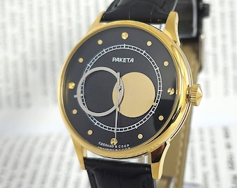 Op de afbeelding: Een gouden polshorloge met een zwarte wijzerplaat en een zwarte leren band. De wijzerplaat van het horloge heeft een uniek ontwerp met een ronde uitsparing en een gouden schijf. De wijzerplaat van het horloge bevat ook de tekst "RAKETA" en "СДЕЛАНО В СССР".