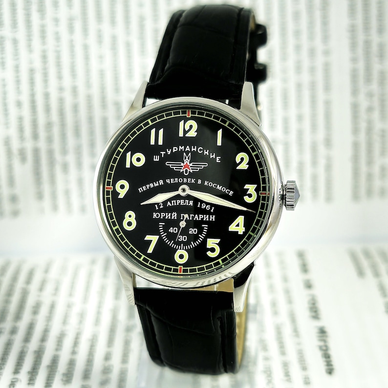 Op de afbeelding: Een zwart polshorloge met een zilveren kast en een zwarte leren band. De wijzerplaat van het horloge is zwart met witte cijfers en een gele secondewijzer. De wijzerplaat van het horloge bevat ook de tekst "ШТУРМАНСКИЕ" en "ПЕРВЫЙ ЧЕЛОВЕК В КОСМОСЕ" in cyrillisch schrift. De wijzerplaat van het horloge bevat ook de tekst "12 АПРЕЛЯ 1961" en "ЮРИЙ ГАГАРИН" in cyrillisch schrift.
