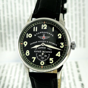 May include: A black and silver wristwatch with a black leather band. The watch face has white numbers and a green outer ring. The watch face also has the text "ШТУРМАНСКИЕ" and "ПЕРВЫЙ ЧЕЛОВЕК В КОСМОСЕ" in Cyrillic script. The watch face also has the text "12 АПРЕЛЯ 1961" and "ЮРИЙ ГАГАРИН" in Cyrillic script.