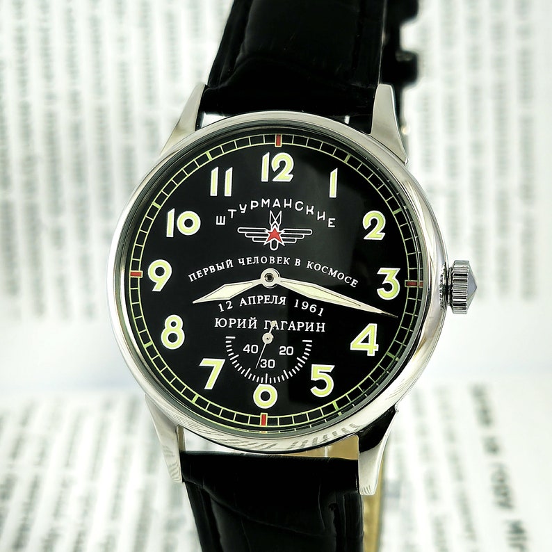 Op de afbeelding: Een zwart-zilveren polshorloge met een zwarte leren band. De wijzerplaat van het horloge heeft witte cijfers en een groene buitenring. De wijzerplaat van het horloge heeft ook de tekst "ШТУРМАНСКИЕ" en "ПЕРВЫЙ ЧЕЛОВЕК В КОСМОСЕ" in cyrillisch schrift. De wijzerplaat van het horloge heeft ook de tekst "12 АПРЕЛЯ 1961" en "ЮРИЙ ГАГАРИН" in cyrillisch schrift.
