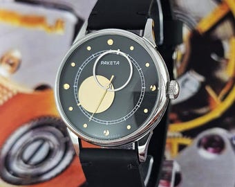 Puede incluir: Un reloj de pulsera plateado con esfera negra y correa de cuero negro. La esfera del reloj presenta detalles dorados y la palabra "PAKETA". El reloj tiene una esfera redonda y una corona texturizada.