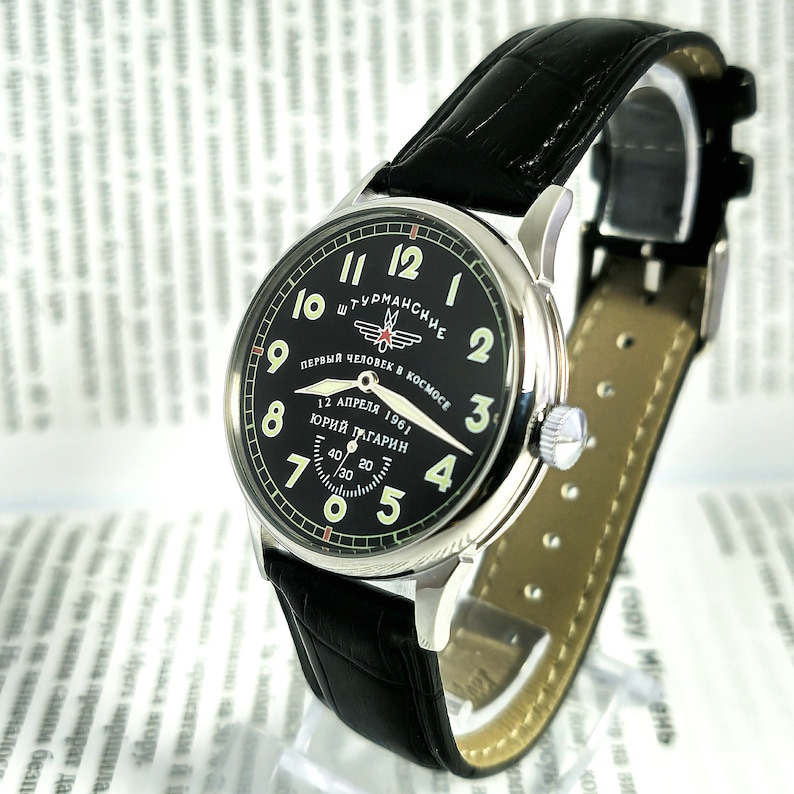 Op de afbeelding: Een zwart en zilveren polshorloge met een zwarte leren band. De wijzerplaat van het horloge heeft een zwarte achtergrond met witte cijfers en een rode ster. De tekst op de wijzerplaat van het horloge luidt "ШТУРМАНСКИЕ ПЕРВЫЙ ЧЕЛОВЕК В КОСМОСЕ 12 АПРЕЛЯ 1961 ЮРИЙ ГАГАРИН".