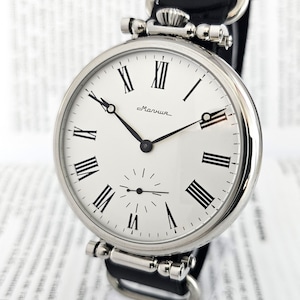 Op de afbeelding: Een zilveren zakhorloge met Romeinse cijfers op een witte wijzerplaat. Het horloge heeft een zwart leren bandje en staat "Машиша" op de wijzerplaat.