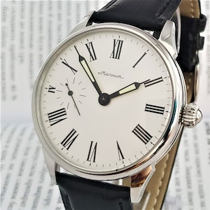 Puede incluir: Un reloj de pulsera plateado con una esfera blanca y marcadores de hora en números romanos negros. El reloj tiene una correa de cuero negra y una pequeña aguja de segundos.