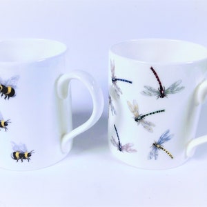 May include: Two white ceramic mugs with handles. One mug features a pattern of bumblebees, the other a pattern of dragonflies.