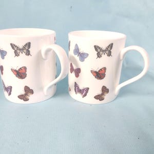 May include: Two white ceramic mugs with curved handles. Each mug is decorated with colourful butterfly illustrations in shades of blue, red, brown, and purple. The mugs are set against a light blue background.