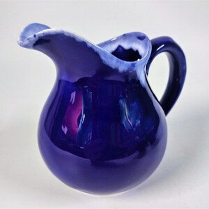 May include: A small, blue ceramic creamer with a white rim and a slightly flared spout. The creamer has a rounded body and a handle.