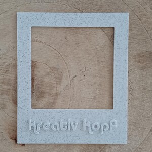Könnte beinhalten: Ein hellgrauer, gesprenkelter, quadratischer Bilderrahmen mit dem geprägten Wort "Kreativ Kopf" am unteren Rand. Der Rahmen befindet sich vor einem hellbraunen Holzgrund.