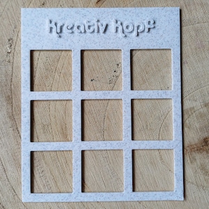 Könnte beinhalten: Ein weißes, rechteckiges Bastelwerkzeug mit neun quadratischen Aussparungen. Der Text "Kreativ Hopp" ist oben eingeprägt. Das Werkzeug besteht aus einem gesprenkelten, hellfarbigen Material und liegt auf einer Holzoberfläche.