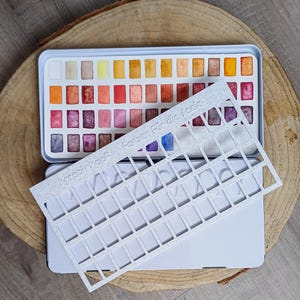 Könnte beinhalten: Ein Aquarellfarbenset in einer rechteckigen Metallbox mit einer Reihe leuchtender Farben. Eine weiße, gitterartige Schablone mit dem Text "kreativ kopf Farben für die Lade" liegt über dem Set. Das Set wird auf einer Holzoberfläche präsentiert.