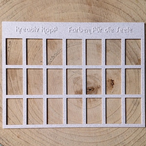 Könnte beinhalten: Eine rechteckige, cremefarbene Schablone mit einem Raster aus rechteckigen Öffnungen. Auf der Schablone stehen die Worte "Kreativ Kopf Farben Für die Seele". Die Schablone liegt auf einer Holzoberfläche.