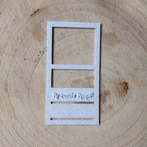 Könnte beinhalten: Ein hellgrauer, rechteckiger Ausstecher mit dem Text "Kreativ Kopp". Der Ausstecher hat oben einen rechteckigen Rahmen, in der Mitte einen horizontalen Balken und unten einen Abschnitt mit zwei horizontalen Linien.