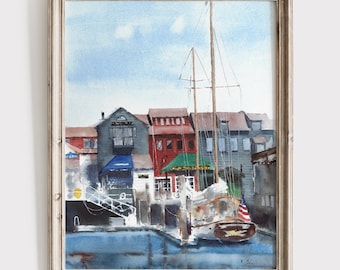 May include: A framed watercolor painting depicting a harbor scene with a sailboat docked in front of colorful buildings. The boat features a small American flag on its stern. The architecture includes red, gray, and blue structures along the water.