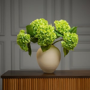 May include: A bouquet of green hydrangeas in a white ceramic vase on a wooden table with a slatted front.