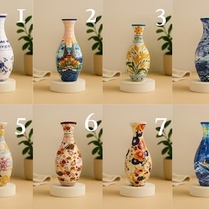 May include: A collection of eight decorative ceramic vases, each with a unique design. The vases vary in shape and color, featuring floral, landscape, and abstract patterns. Each vase is displayed on a small, round, white pedestal.