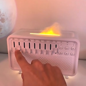 Könnte beinhalten: Eine weiße Soundmaschine mit Knöpfen und Schiebereglern. Sie projiziert ein warmes, gelbes Licht. Ein Finger drückt einen Schieberegler. Die Maschine hat einen Lautsprecher und Symbole. Sie ist für den Schlaf konzipiert.