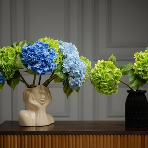 May include: Two arrangements of artificial hydrangeas in vases. One vase is a white ceramic sculpture of a face, the other is a black textured vase. Both vases are on a wooden table with a bamboo-like pattern.