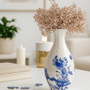 May include: A white puzzle vase with blue floral designs holds dried flowers. Several puzzle pieces are scattered on the table. A candle and a stack of books are also visible.