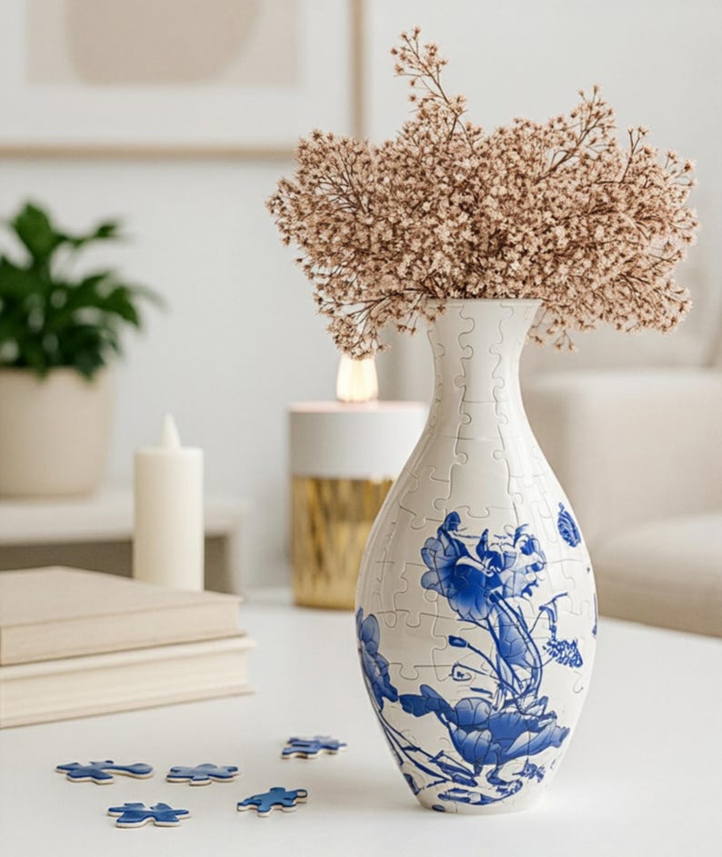May include: A white puzzle vase with blue floral designs holds dried flowers. Several puzzle pieces are scattered on the table. A candle and a stack of books are also visible.