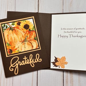 May include: A dark brown Thanksgiving greeting card featuring a watercolor illustration of orange pumpkins and yellow sunflowers on the front with the word Grateful in script. The inside contains the text In this season of gratitude, I'm thankful for you. Happy Thanksgiving and a small paper maple leaf.