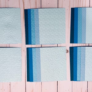 May include: Six sets of square cardstock paper in shades of blue, ranging from dark navy to light sky blue. Each set features a distinct embossed texture, including floral patterns, geometric grids, polka dots, script, and swirling designs.