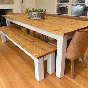 May include: A rectangular dining table with a natural wood grain top and white painted legs, accompanied by a matching wooden bench and several brown upholstered chairs. A woven basket containing green plants sits on the centre of the table.