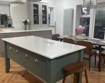 Puede incluir: Una isla de cocina con base verde oscuro, encimera blanca y tiradores de latón, acompañada de un taburete de madera. La habitación cuenta con paredes blancas, suelo de madera en espiga, tres lámparas colgantes blancas y un comedor con sillas.