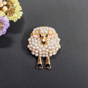 May include: A gold-coloured sheep brooch with a pearl-like texture. The sheep has pink ears, a black face, and black hooves. The brooch is a decorative accessory.