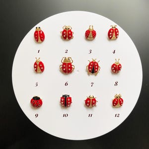 May include: A collection of twelve ladybug-themed brooches. Each brooch features a red body with black spots and gold-colored metal accents. The ladybugs vary in design, with some featuring crystal embellishments.