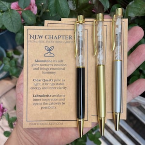 Könnte beinhalten: Drei Stifte mit goldenen Akzenten und transparenten Schäften, gefüllt mit kleinen Steinen. Ein Stift hat einen schwarzen Schaft, einer ist goldfarben und einer silberfarben. Eine Karte hinter den Stiften lautet "NEW CHAPTER" und listet die Eigenschaften der Steine auf.